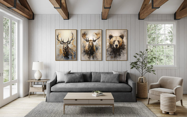 Set of 3 framed wildlife paintings of an elk, moose, and bear hanging above a gray sofa in a cabin.
