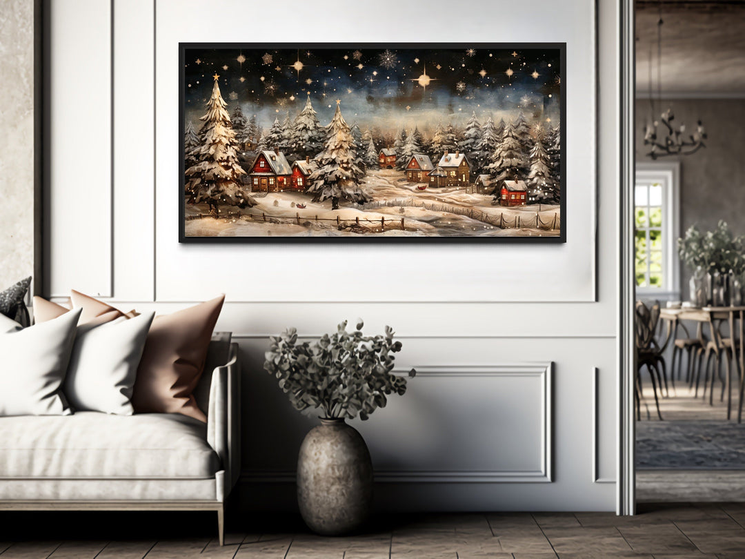Snowy winter village night scene wall art in a black frame above a sofa in a modern living room.