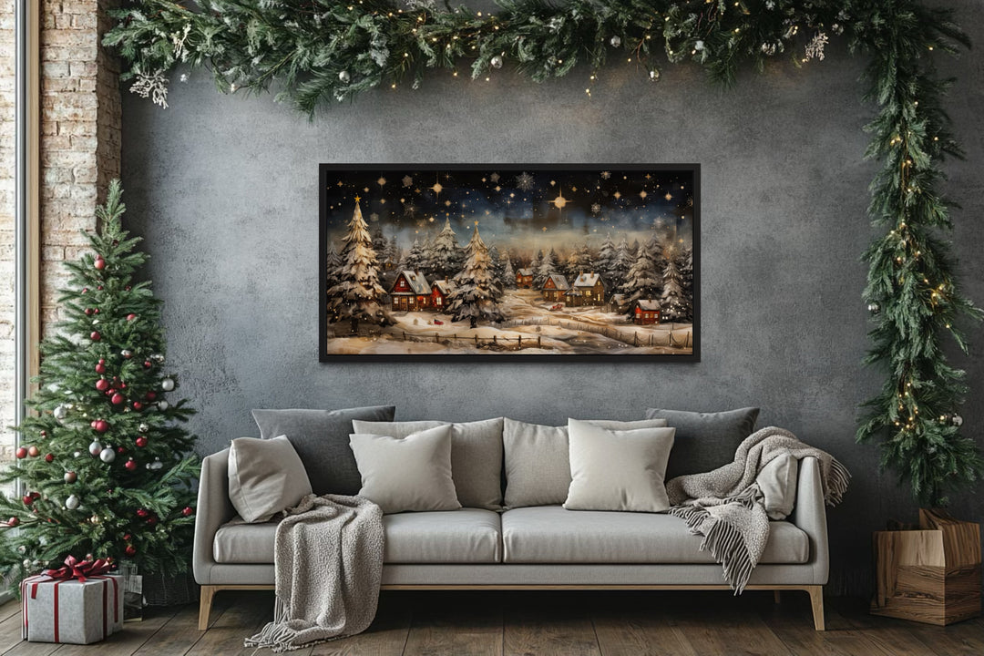 Snowy winter village at night wall art in a black frame above a sofa in a Christmas-themed room.