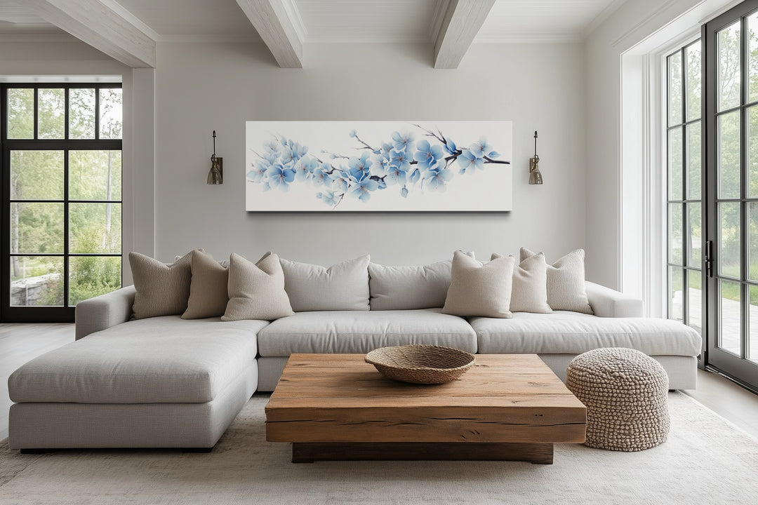 Long horizontal wall art of soft blue blossom flowers hanging above a neutral sofa in a living room.