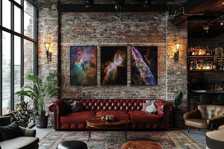 Three vertical space nebula canvas prints displayed above a red leather sofa in a brick loft.