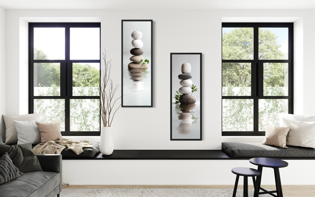 Two tall, narrow canvas prints of stacked zen stones hanging on a white wall between two windows.