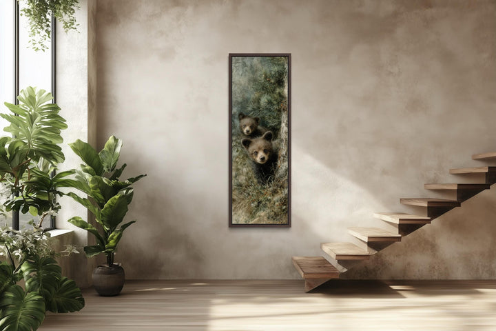 Tall narrow canvas wall art of two baby bear cubs peeking from a pine tree in a rustic room.