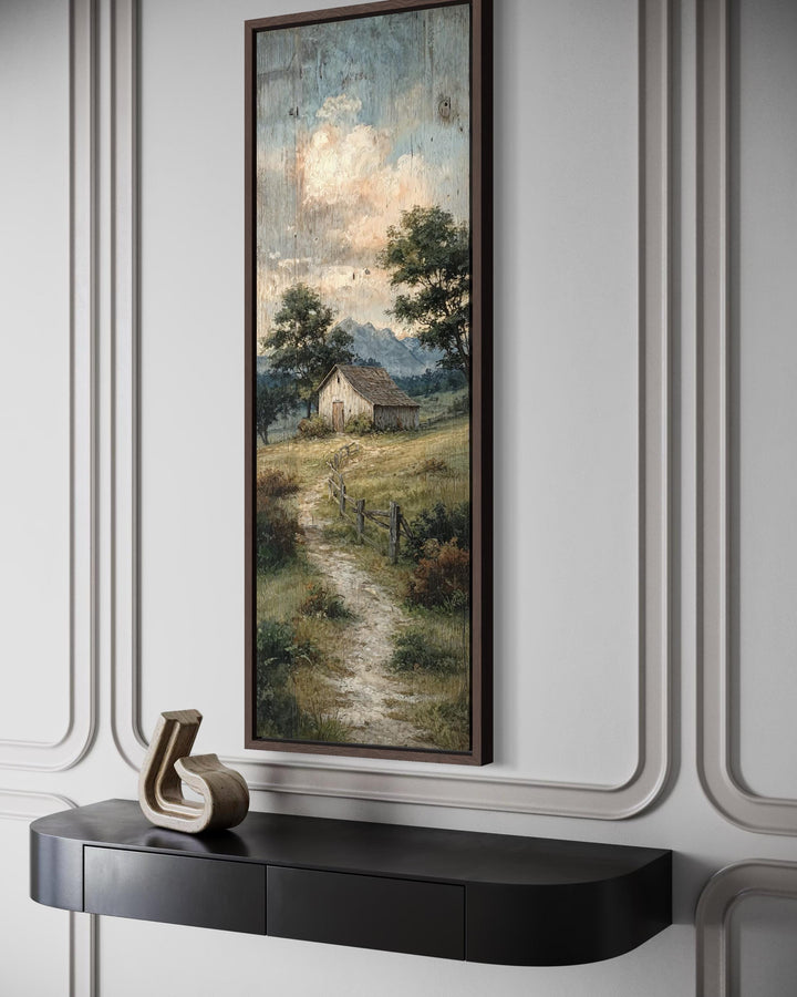 Tall narrow framed canvas art of a rustic barn and country path landscape on a paneled wall.