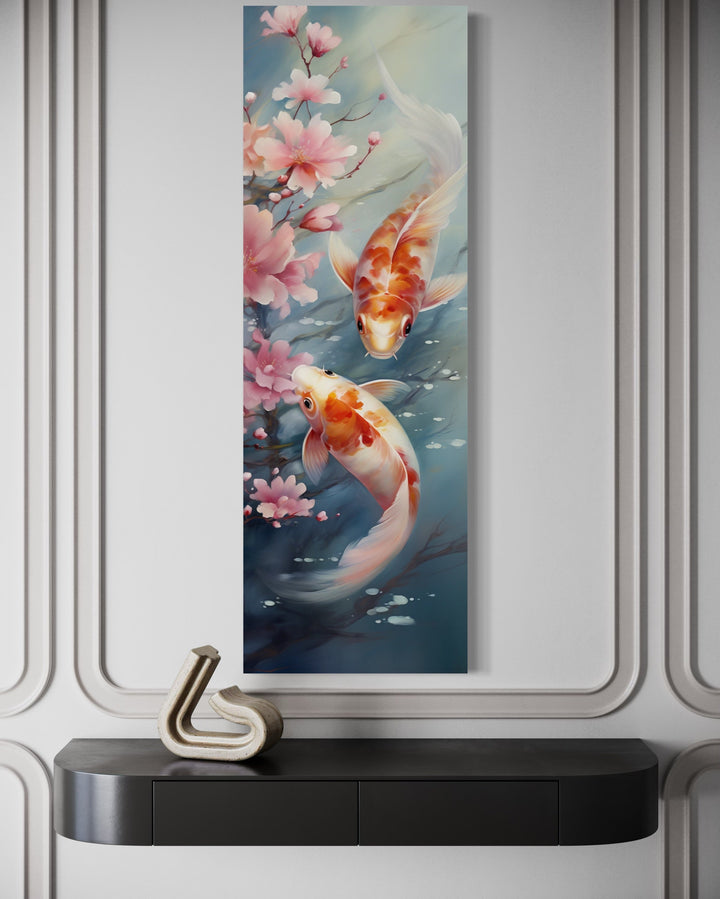 Tall narrow vertical canvas art of two koi fish swimming among pink cherry blossoms on a white wall.