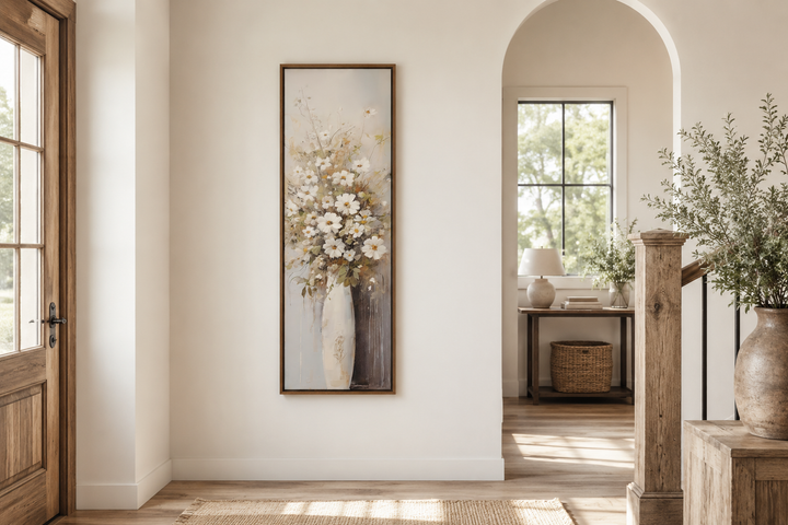Tall narrow vertical framed canvas art of white wildflowers in a vase on a neutral wall.