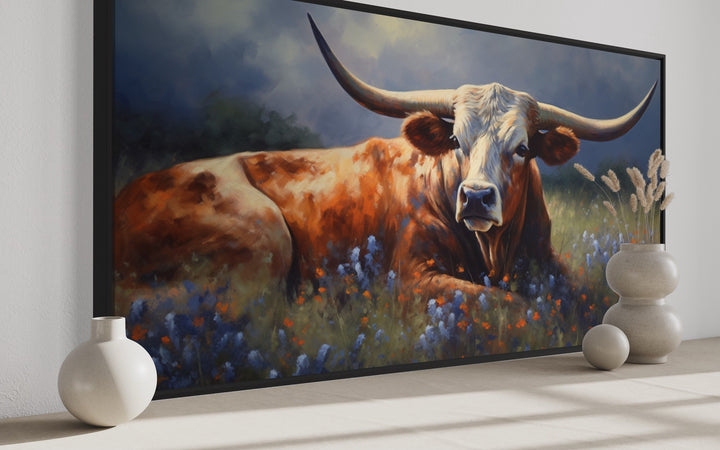 Framed canvas print of a Texas Longhorn cow lying in a field of bluebonnets and wildflowers.