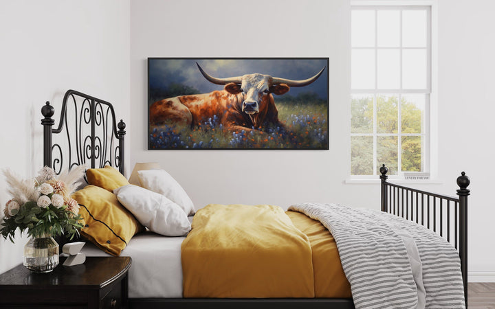Framed canvas print of a Texas Longhorn cow in a bluebonnet field above a bed in a rustic bedroom.