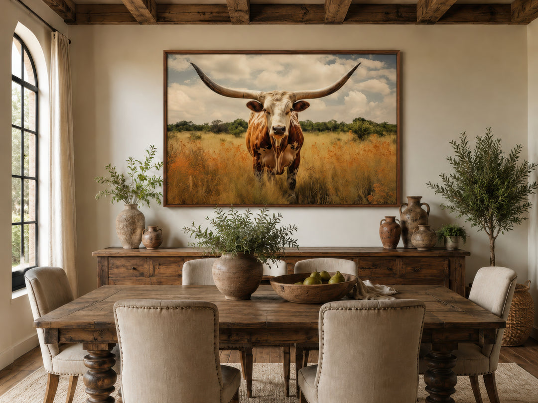 Framed canvas wall art of a Texas Longhorn cow in a field above a rustic wooden dining table.