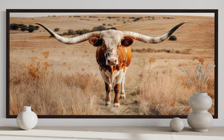 Framed canvas wall art of a Texas longhorn bull in a golden ranch landscape above a white shelf.