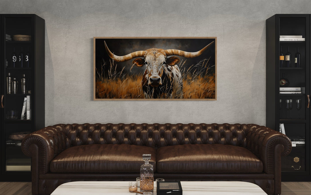 Framed canvas painting of a Texas longhorn steer in tall grass above a brown leather sofa.