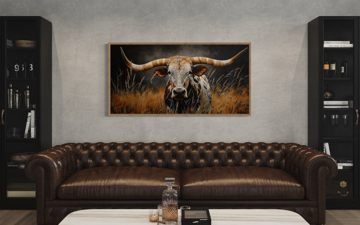 Framed canvas painting of a Texas longhorn steer in tall grass above a brown leather sofa.