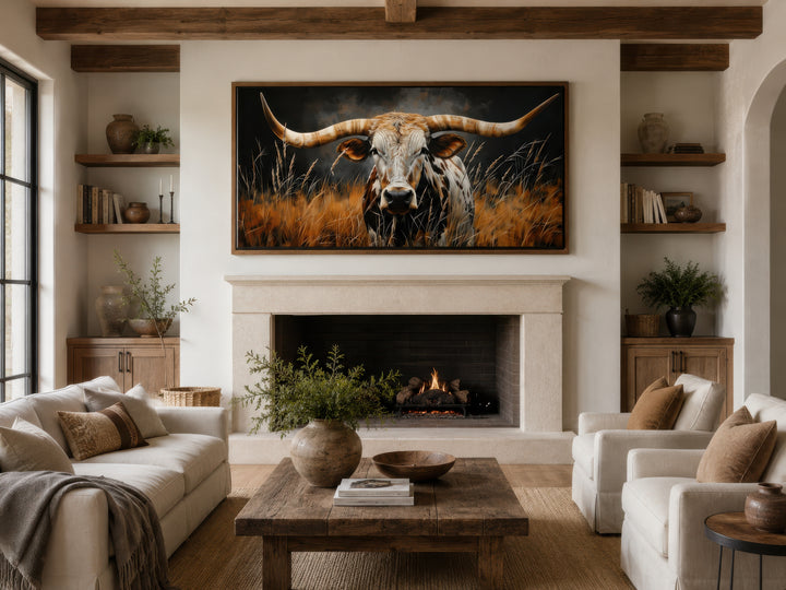 Framed canvas wall art of a Texas longhorn steer in grass, hung above a fireplace in a rustic room.