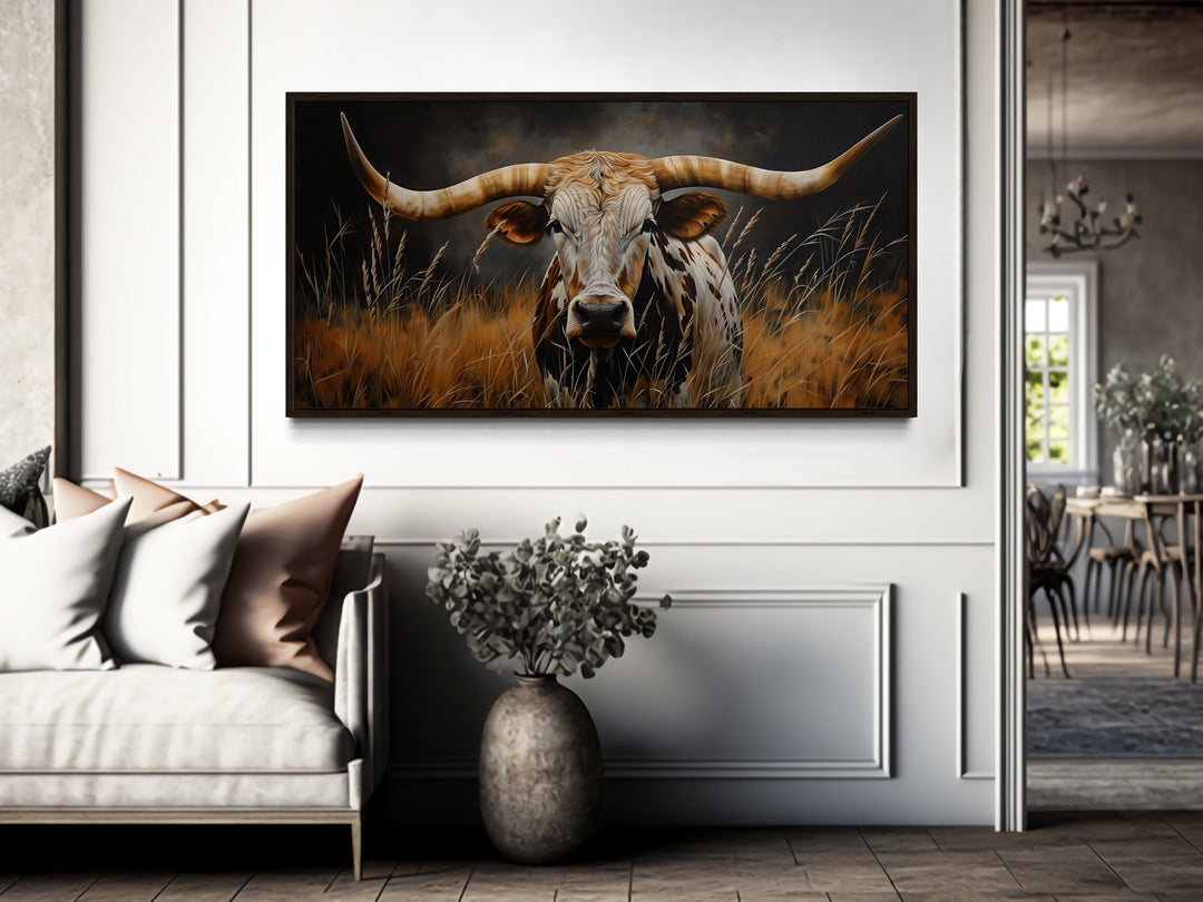 Framed canvas wall art of a Texas longhorn steer in golden grass above a sofa in a rustic room.