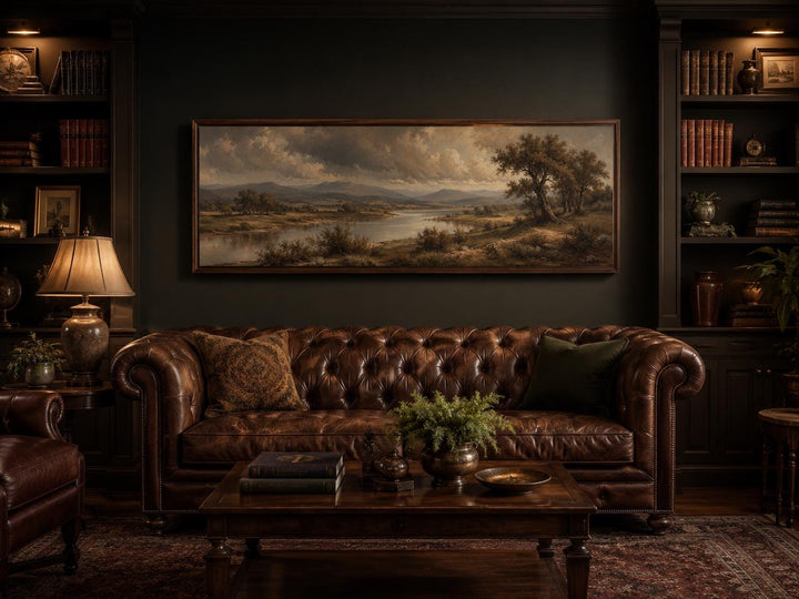 Long horizontal canvas print of a moody river valley landscape above a brown leather sofa.