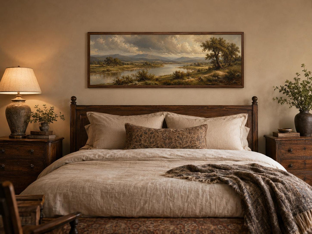 Traditional landscape canvas of a moody river valley above a bed with dark wood furniture.