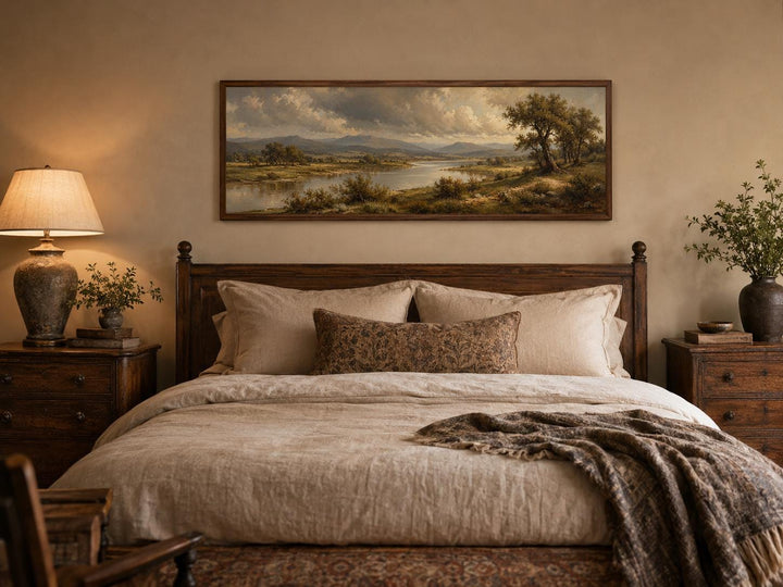 Traditional landscape canvas of a moody river valley above a bed with dark wood furniture.