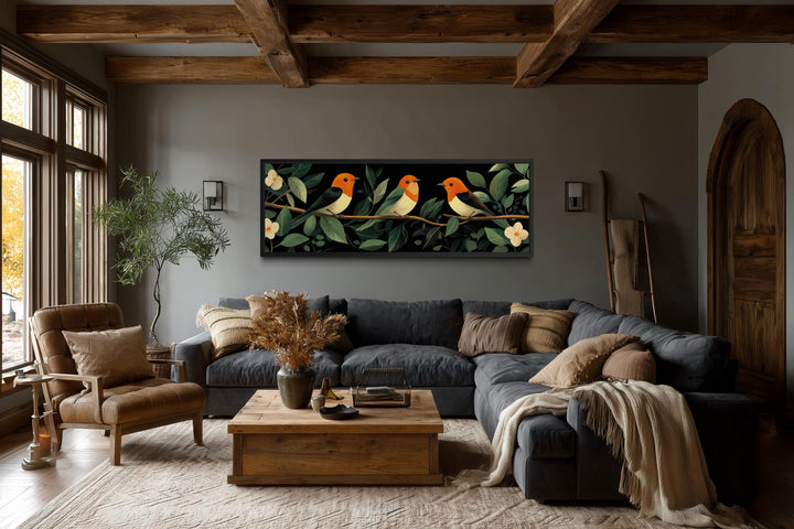 Panoramic canvas print of three orange-headed tropical birds on a branch over a dark grey sofa.
