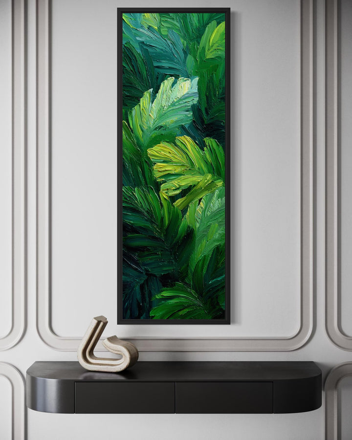 Vertical canvas print of textured green tropical leaves in a black frame above a dark console table.