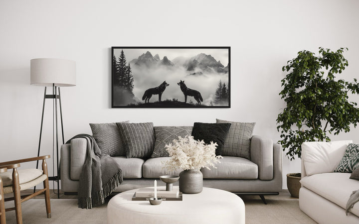 Black and white framed canvas art of two wolves on a cliff in foggy mountains above a gray sofa.