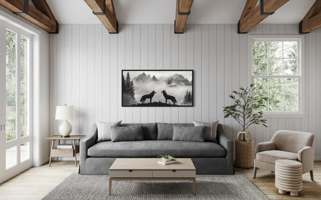 Black and white framed canvas of two wolves on a cliff in foggy mountains above a gray sofa.
