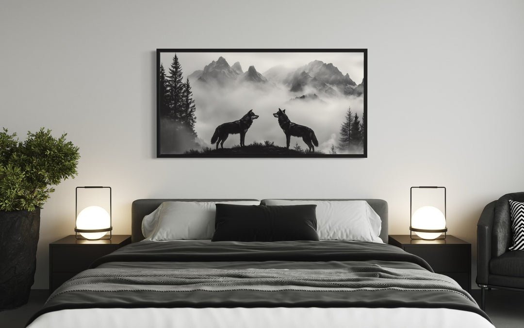 Two wolves on a cliff in foggy mountains black and white framed canvas art above a bed.