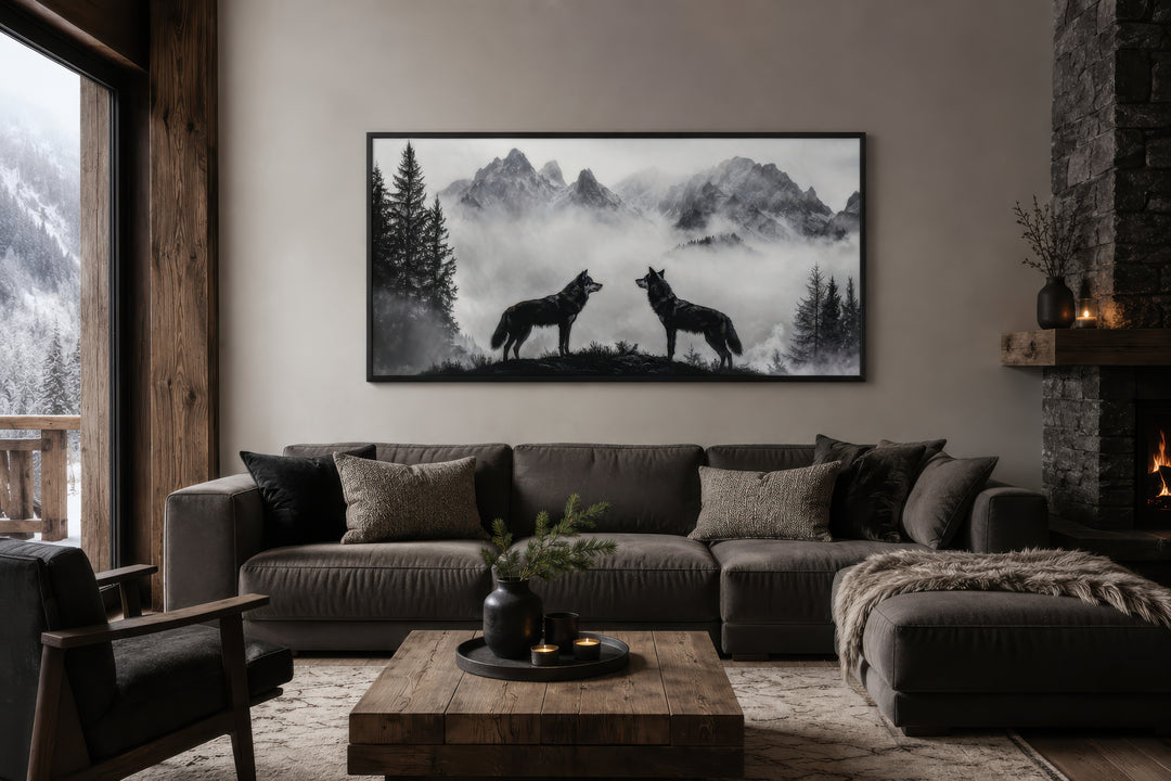 Black and white framed canvas wall art of two wolves on a cliff in foggy mountains above a sofa.