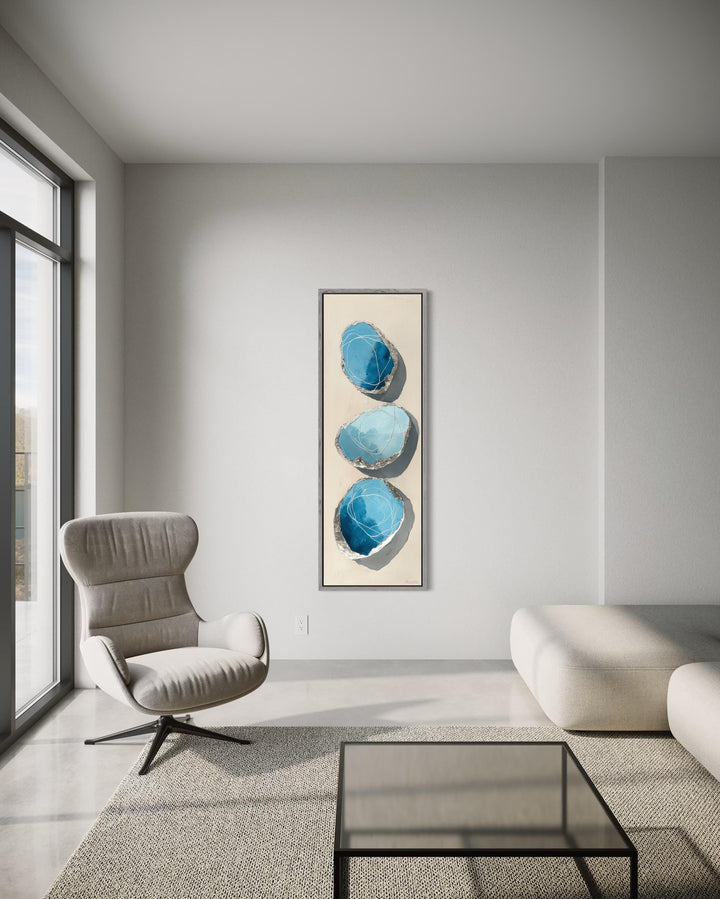 Vertical tall narrow canvas print featuring three abstract blue and silver stones on a neutral wall.