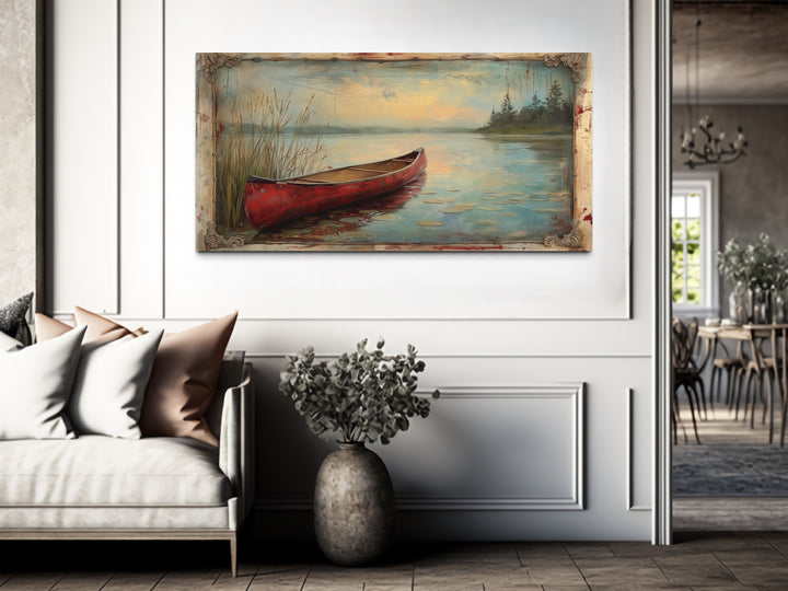 Vintage red canoe on a calm lake painting in a rustic frame hanging above a sofa in a cabin.
