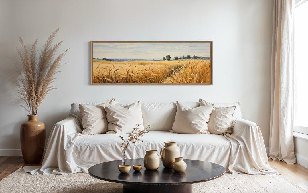 Long horizontal framed canvas wall art of a golden wheat field landscape above a white sofa.