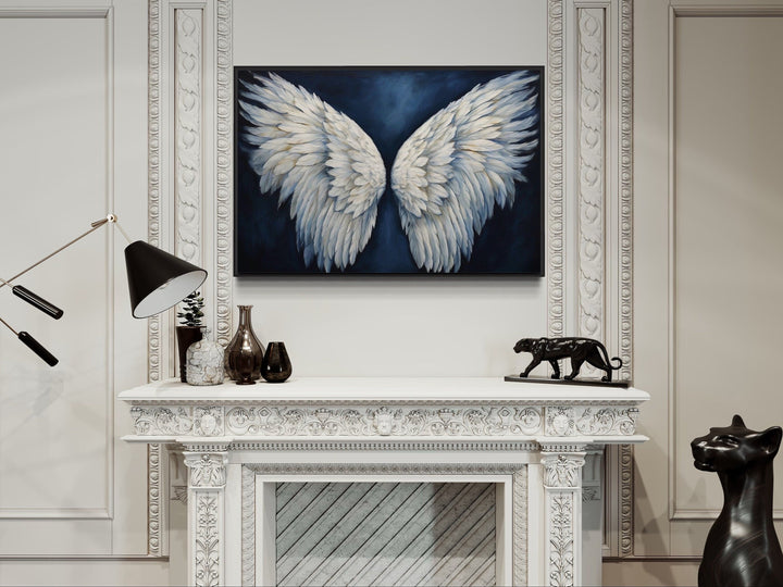 White angel wings on a dark blue background framed canvas wall art above a white fireplace mantel.