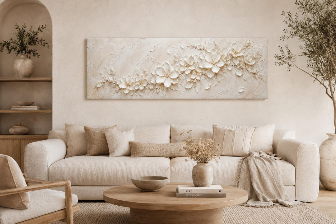 Long horizontal white floral canvas art with textured ivory blossoms above a neutral sofa.