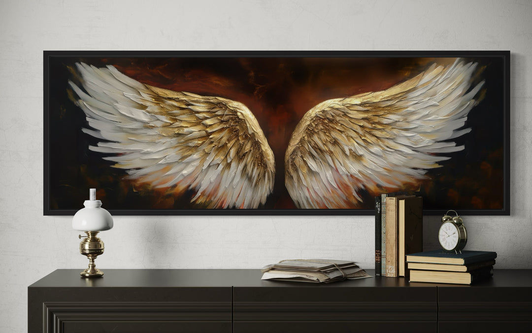 Horizontal framed canvas of white and gold angel wings on a dark brown background above a dresser.