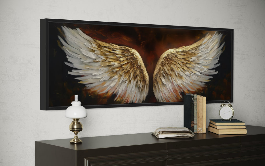 Horizontal framed canvas of white and gold angel wings on a brown background above a dark dresser.
