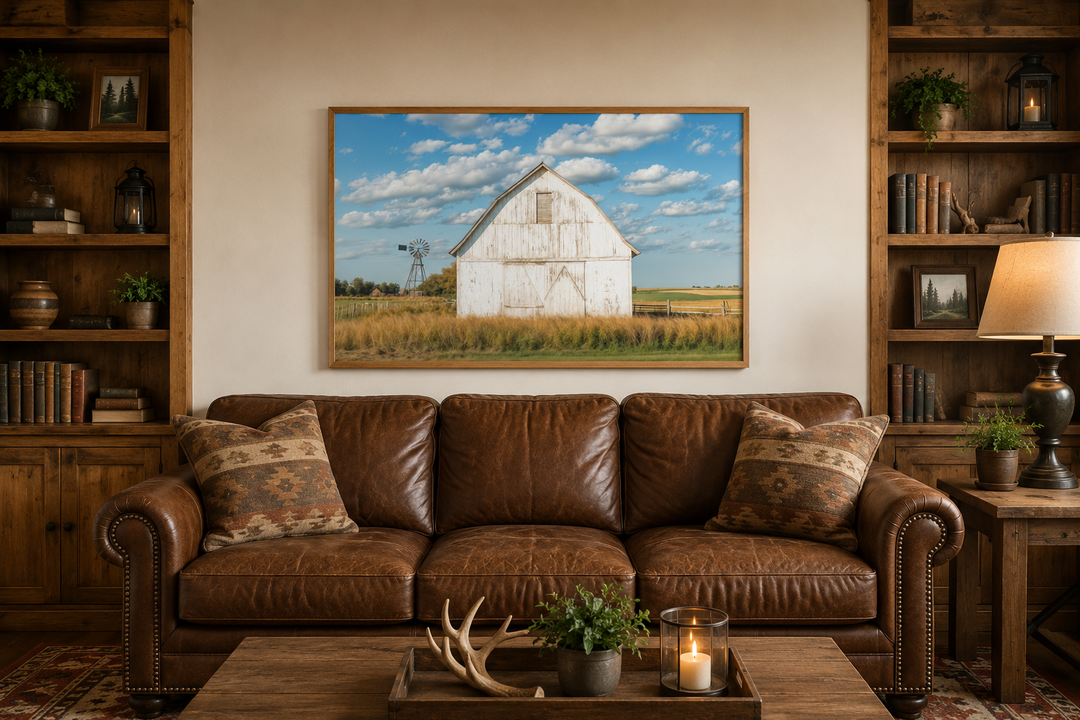 Framed canvas wall art of a weathered white barn and windmill above a brown leather sofa.