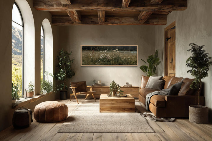 Horizontal wildflower meadow canvas art in a light wood frame above a modern rustic living room sofa.