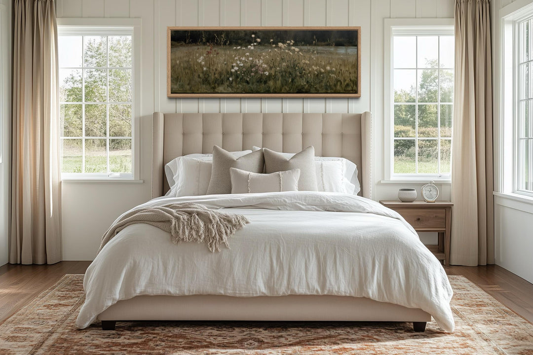 A long, narrow wildflower meadow landscape canvas in a light wood frame hangs above a neutral bed.
