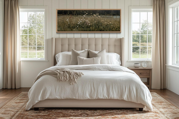 A long, narrow wildflower meadow landscape canvas in a light wood frame hangs above a neutral bed.