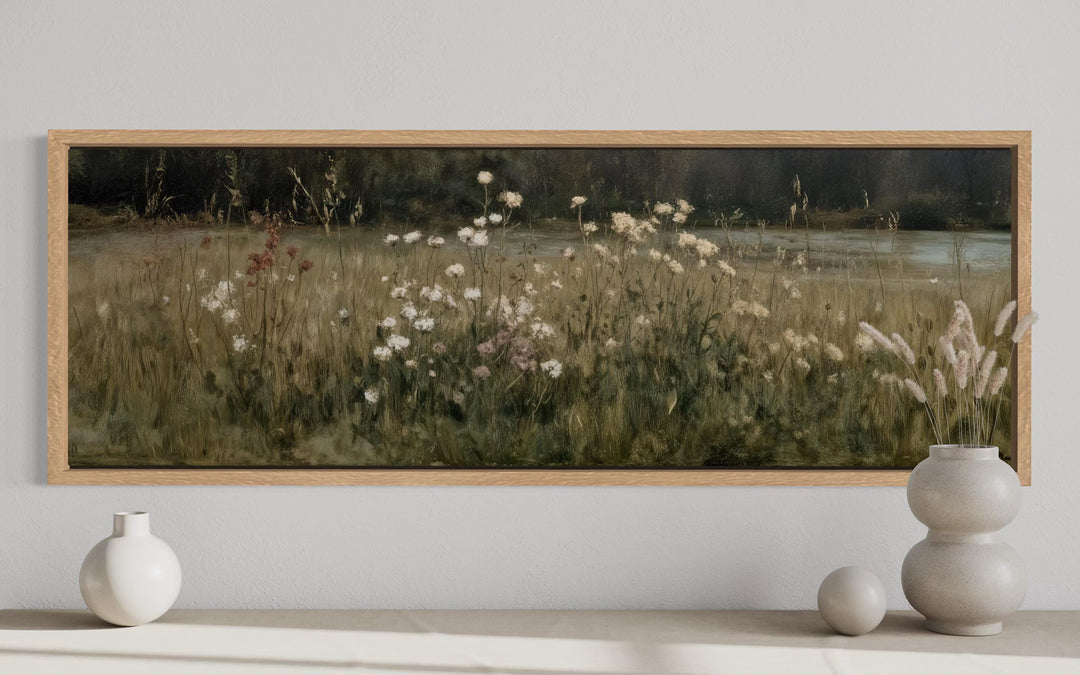 A long, narrow canvas print of a wildflower meadow in a light wood frame above a console table.