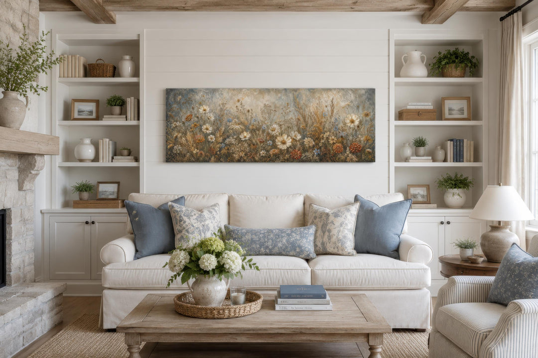 A long narrow canvas print of a neutral wildflower meadow hangs above a white sofa in a cozy room.