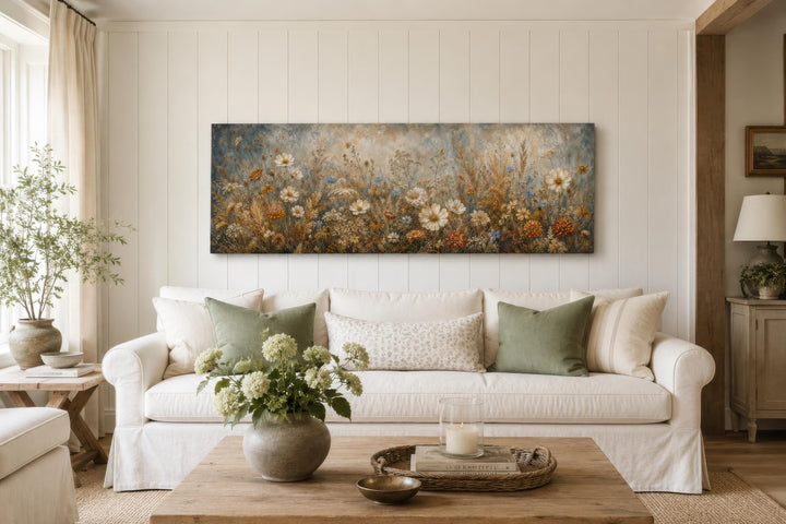 A long narrow canvas print of neutral wildflowers hangs above a white sofa in a bright living room.