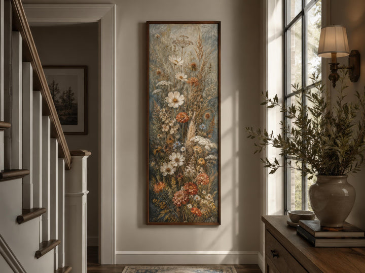 Tall vertical canvas print of blue, rust, and cream wildflowers in a dark wood frame on a tan wall.