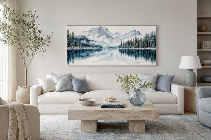 Framed horizontal canvas print of a winter mountain lake and pine forest above a white sofa.