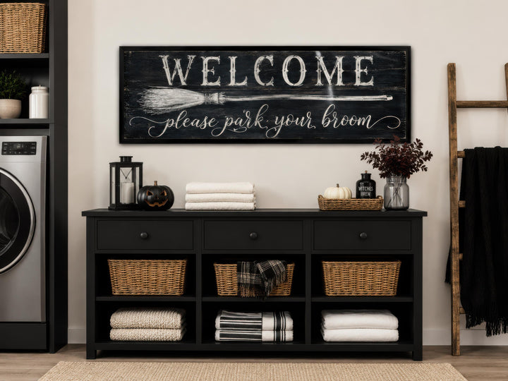 A horizontal black wooden sign with white text reading 'WELCOME please park your broom'.