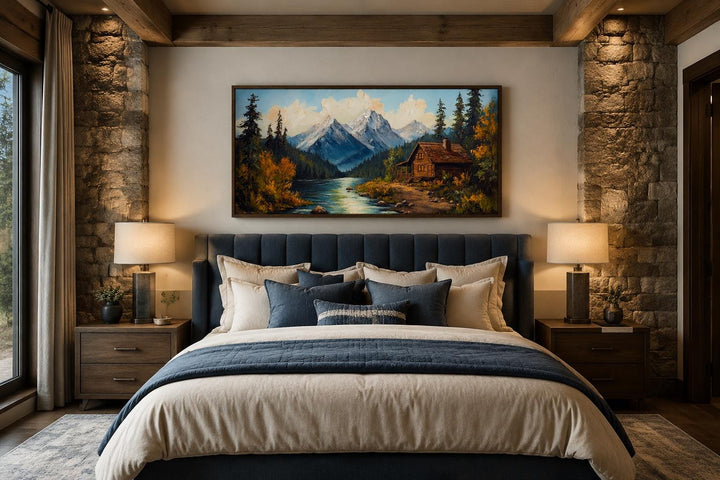 Vintage style canvas wall art of a mountain cabin by a river, hung above a bed in a rustic room.