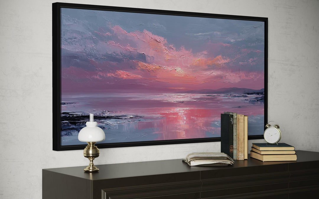 A framed canvas print of a blush pink ocean sunset hangs above a dark wood dresser with decor.