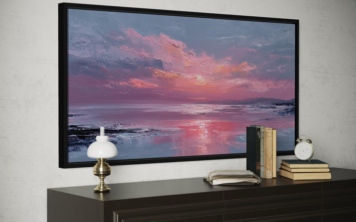 A framed canvas print of a blush pink ocean sunset hangs above a dark wood dresser with decor.
