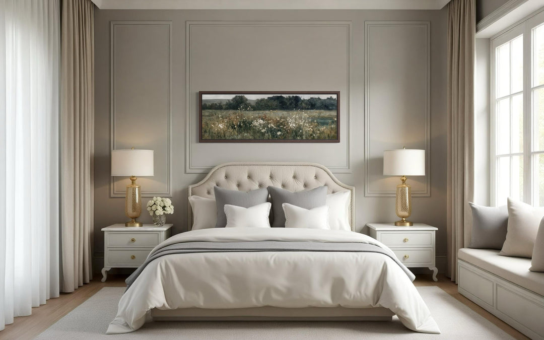 A long narrow horizontal painting of a moody wildflower field hangs above a bed in a neutral room.